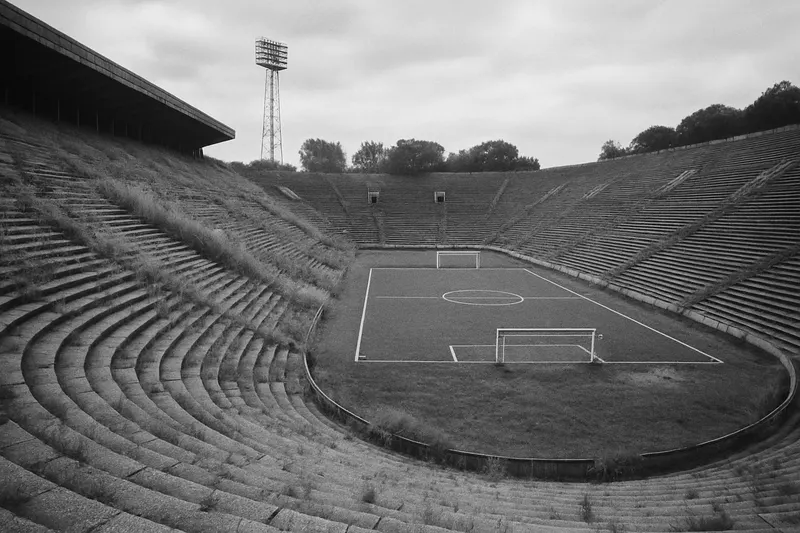 Prázdné betonové tribuny obrovského stadionu porostlé trávou, dole uprostřed malé fotbalové hřiště